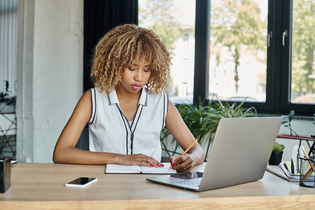 Konsantre Afro-Amerikan iş kadını şık bir ofiste dizüstü bilgisayarın yanında notlar alıyor. - Fotoğraf, Görsel