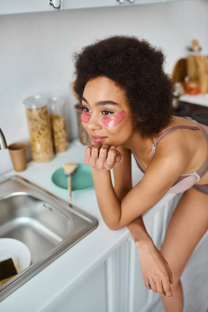 mujer afroamericana complacida en sujetador con parches rosados bajo los ojos sonriendo en la cocina - Foto, Imagen