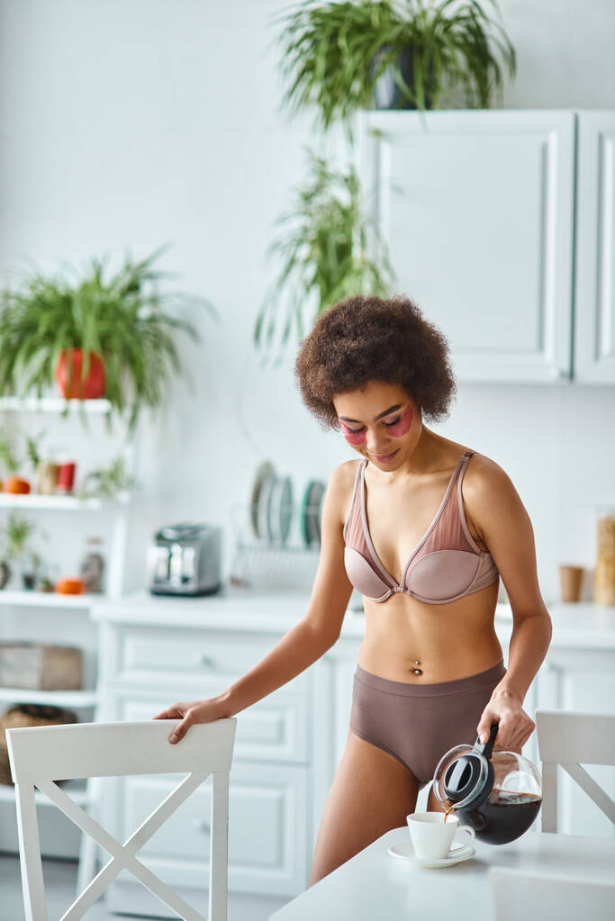 radiante mujer afroamericana con parches para los ojos vertiendo café en taza de porcelana en la cocina - Foto, imagen
