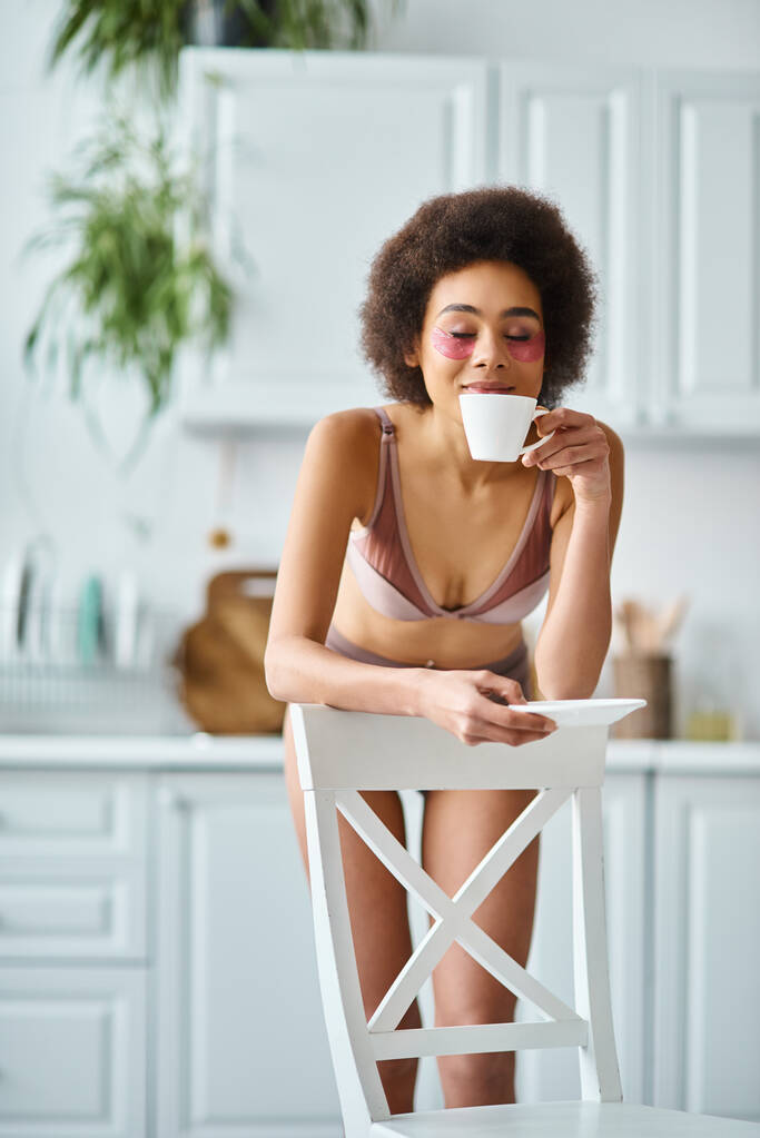 Χαρούμενη Αφροαμερικανή με μπαλώματα στα μάτια που στέκεται με εσώρουχα και μυρίζει πρωινό καφέ. - Φωτογραφία, εικόνα