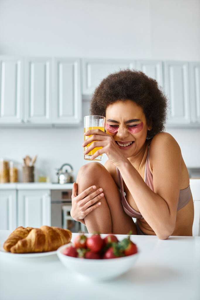 mujer afroamericana feliz en lencería bebiendo jugo de naranja cerca del desayuno en la mesa de la cocina - Foto, Imagen