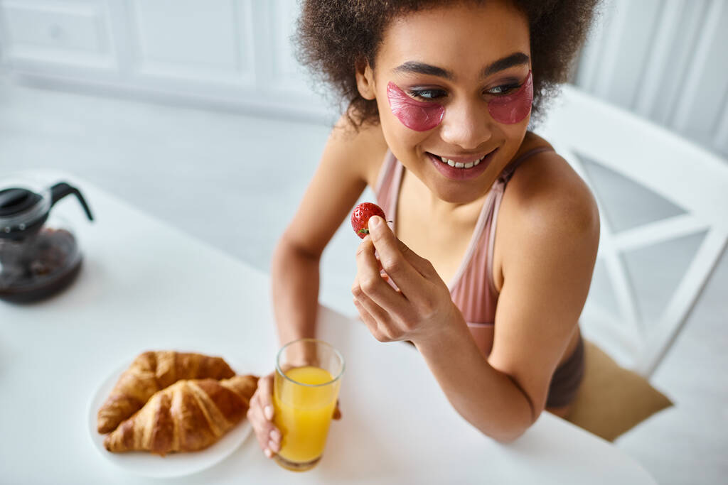heureuse femme afro-américaine manger tenant fraise et verre de jus d'orange dans la cuisine - Photo, image