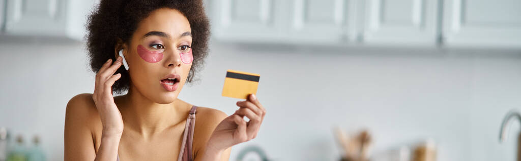 sztandar zaskoczony czarny kobieta w słuchawki z łaty pod oczami patrząc na kartę kredytową - Zdjęcie, obraz