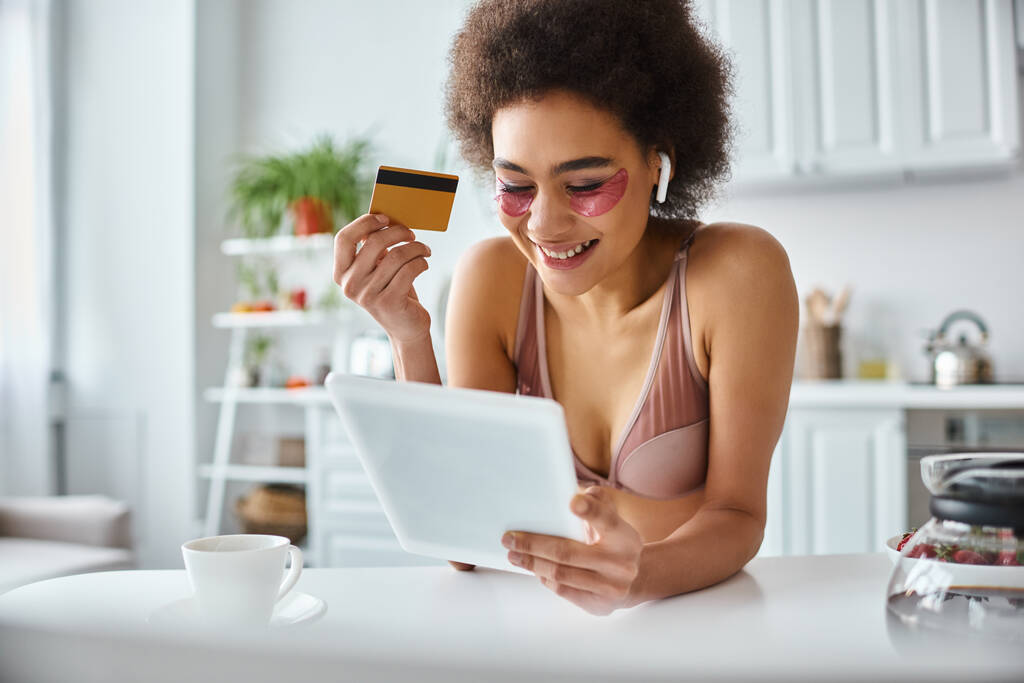 χαρούμενη Αφροαμερικανή γυναίκα που ψωνίζει online με tablet με πιστωτική κάρτα στο χέρι και καφέ στην άκρη - Φωτογραφία, εικόνα