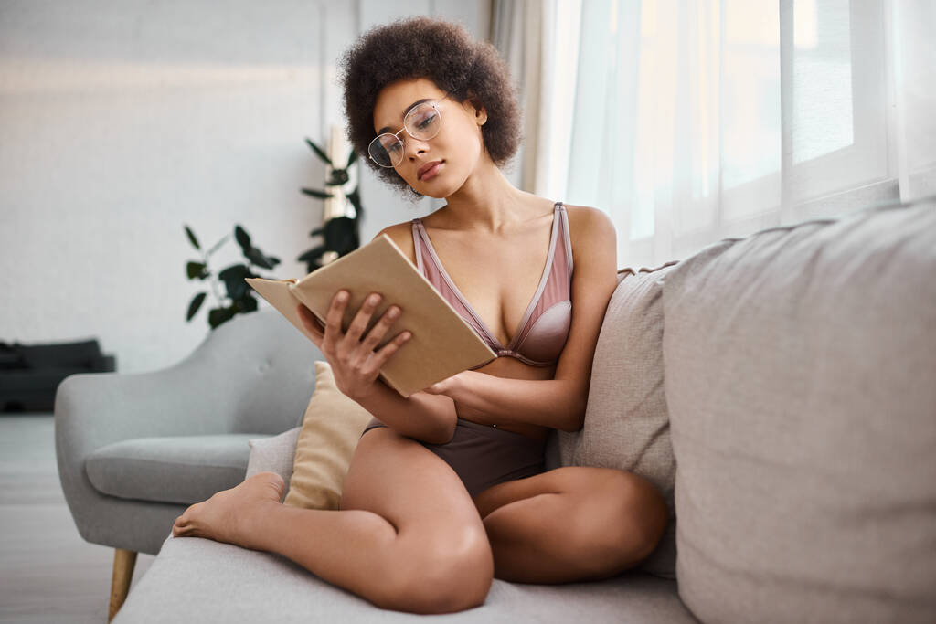 χαλαρή και σγουρά μαλλιά αφροαμερικανή γυναίκα διαβάζοντας ένα βιβλίο με εσώρουχα σε άνετο καναπέ, γαλήνια - Φωτογραφία, εικόνα