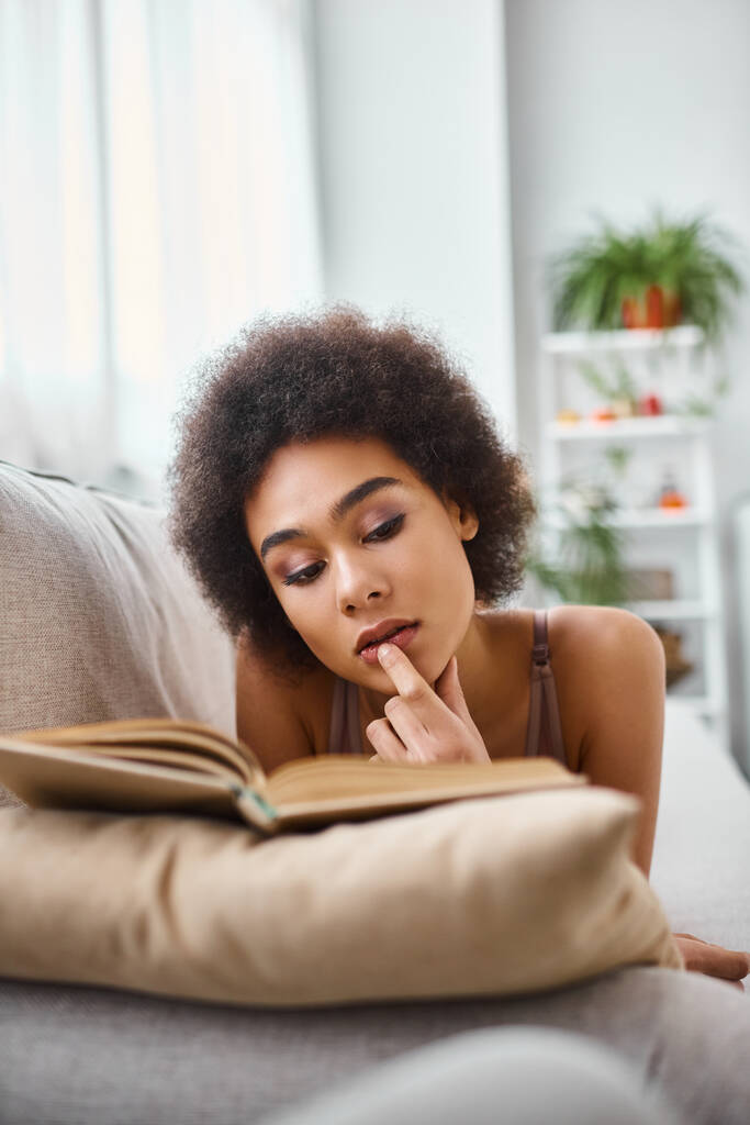 koncentrált és fiatal afro-amerikai nő olvas egy könyvet fehérnemű egy kényelmes kanapén - Fotó, kép