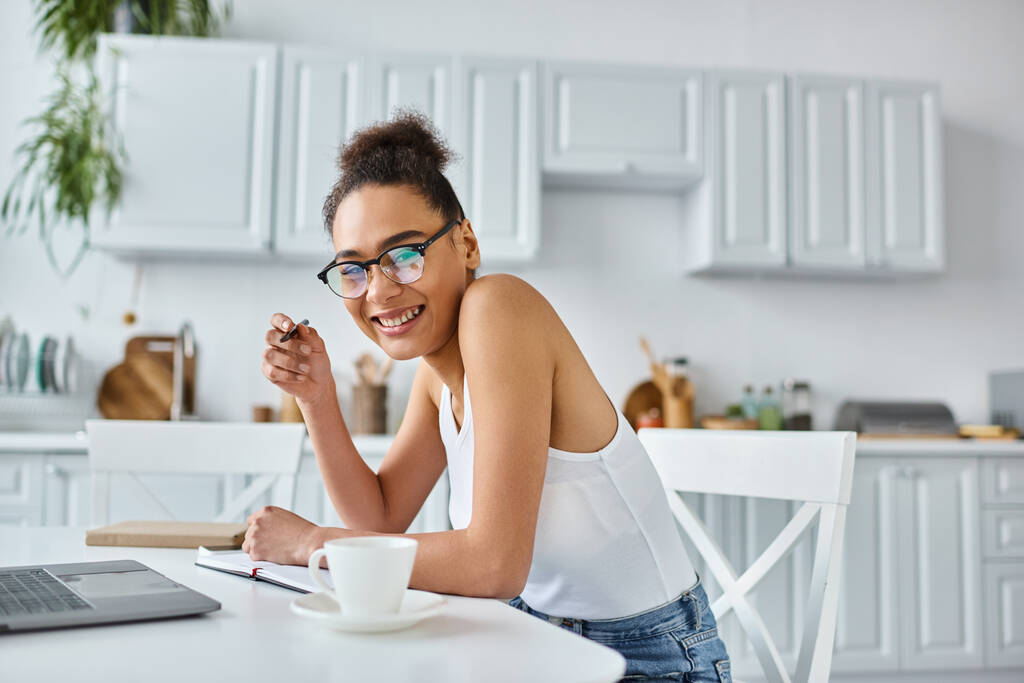 allegra donna afroamericana in occhiali prendere appunti vicino laptop e tazza di caffè, lavoro a distanza - Foto, immagini