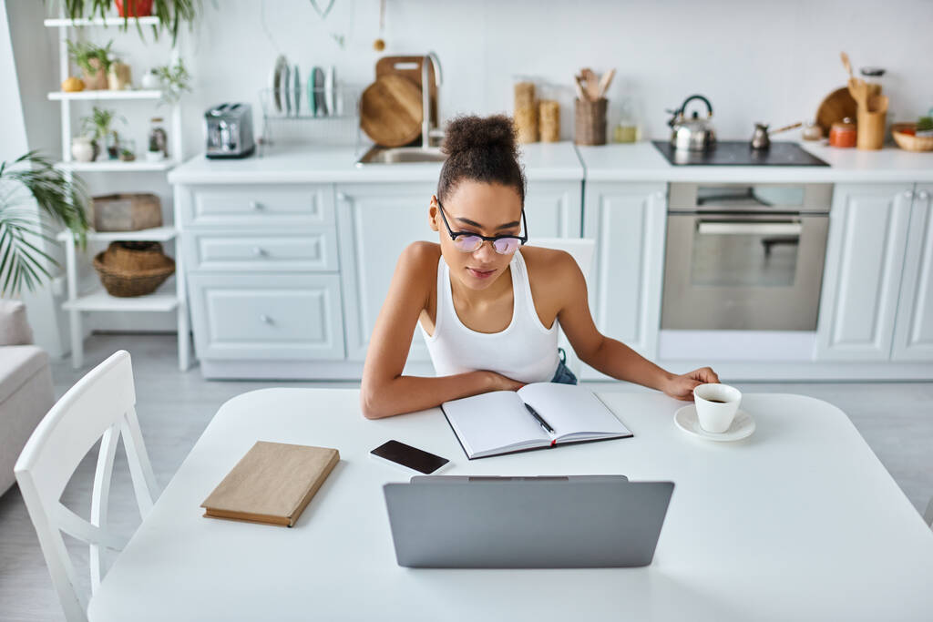 молодая африканская американка в очках смотрит на свой ноутбук возле чашки кофе, удаленная работа - Фото, изображение
