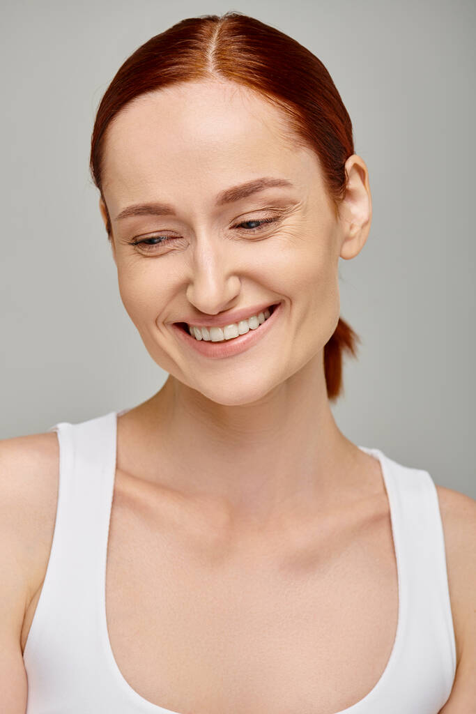 mujer pelirroja feliz en camiseta sin mangas exudando sonrisa alegre y saludable sobre fondo gris, mirando hacia abajo - Foto, Imagen