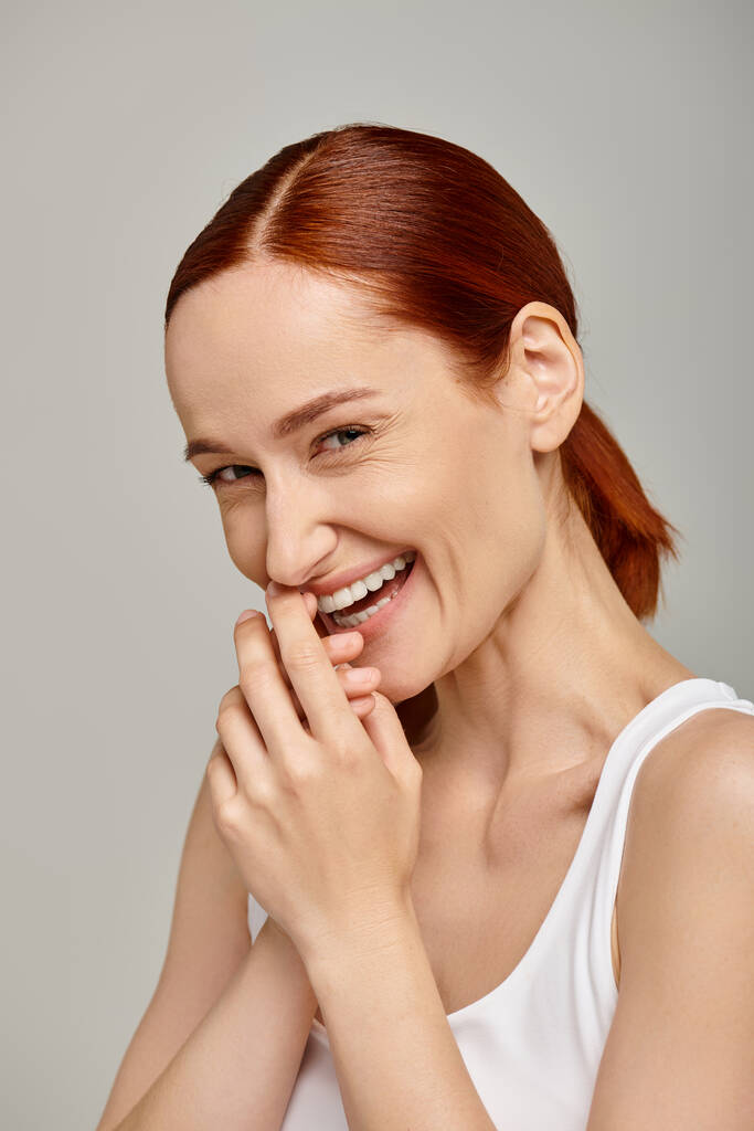 happy and redhead woman in white tank top laughing and looking at camera on grey background - Photo, Image