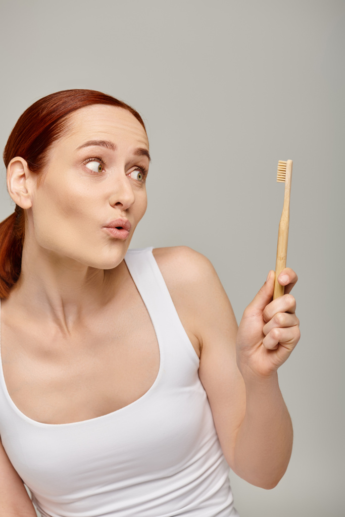 surprised redhead woman in tank top looking at toothbrush on grey background, dental hygiene concept - Photo, Image