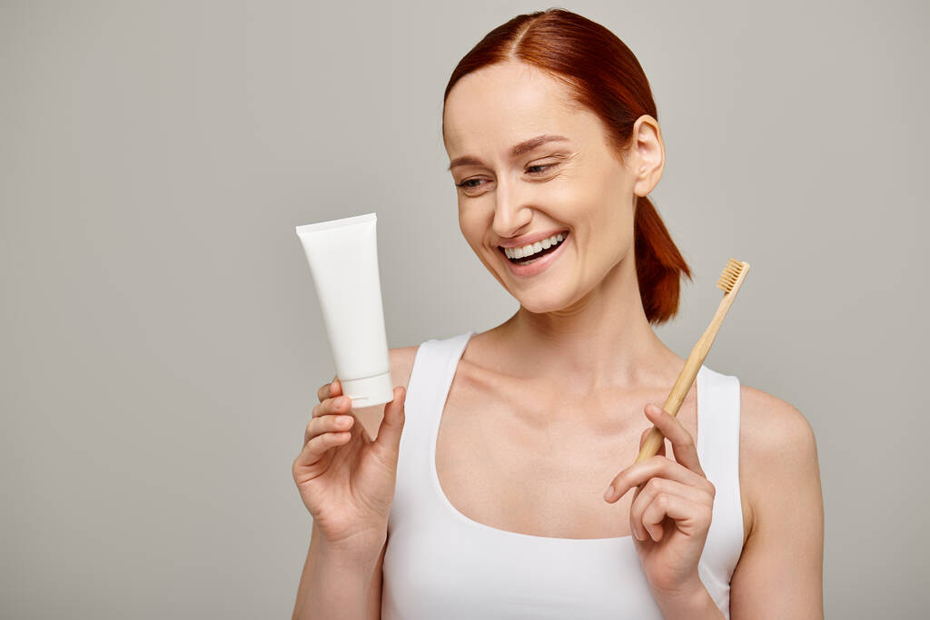 gaie rousse femme en débardeur tenant dentifrice et brosse à dents et souriant à la caméra sur gris - Photo, image