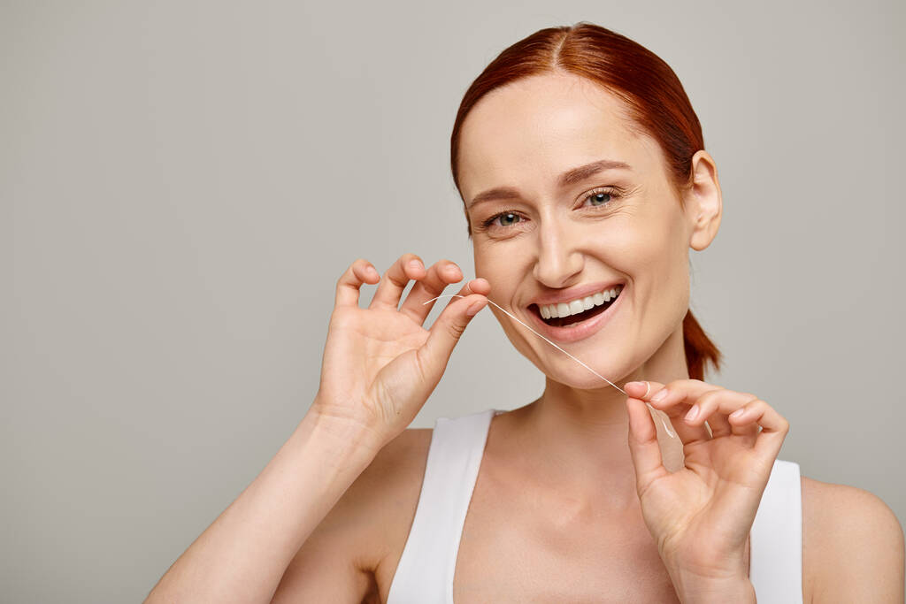 χαρούμενη κοκκινομάλλα γυναίκα που κρατάει οδοντικό νήμα και χαμογελά σε γκρι φόντο, προωθώντας την στοματική υγιεινή - Φωτογραφία, εικόνα