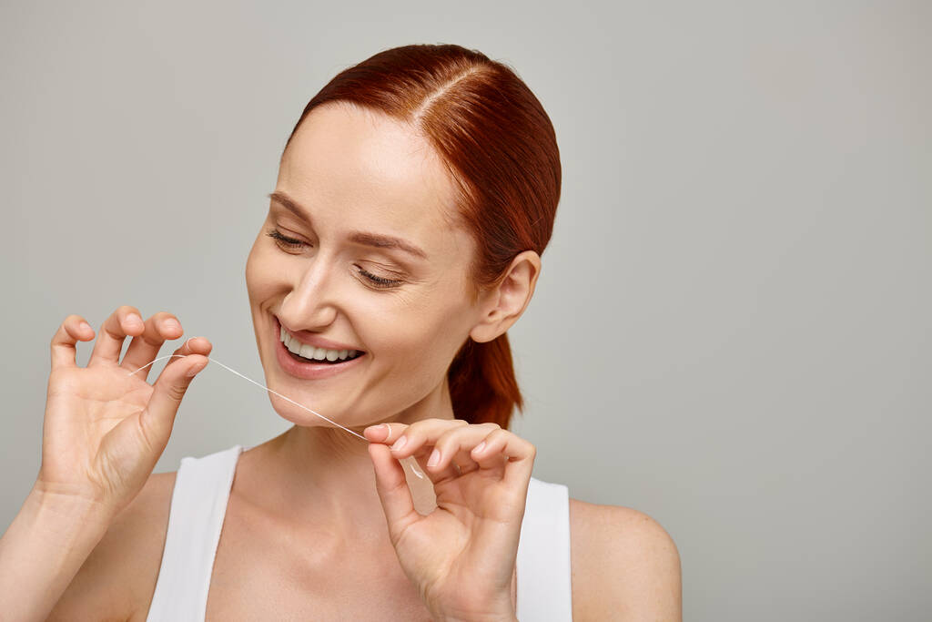 mulher ruiva alegre segurando fio dental e sorrindo em fundo cinza, promovendo a higiene oral - Foto, Imagem