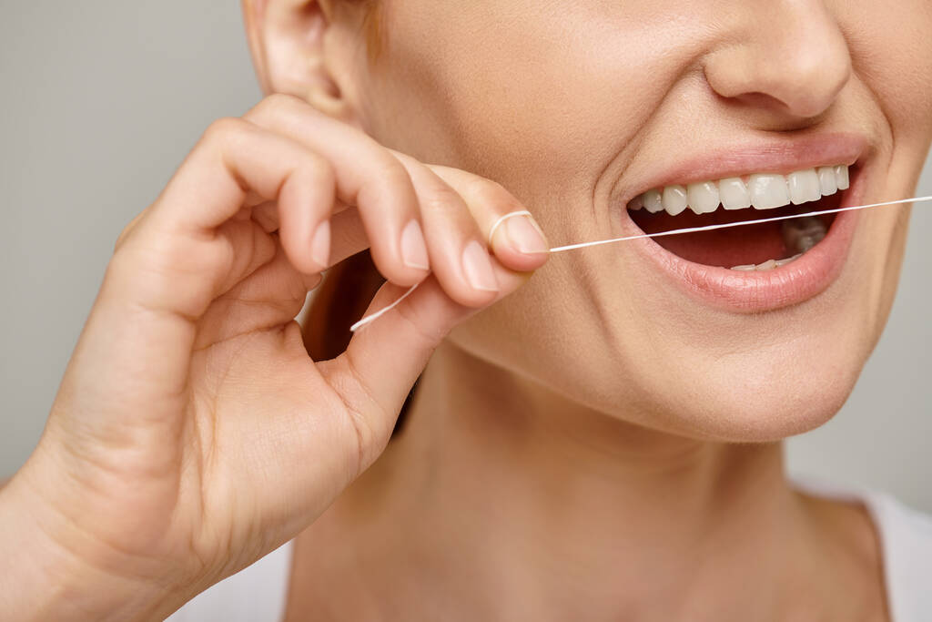 przycięta kobieta trzyma nitkę dentystyczną i uśmiecha się na szarym tle, promując higienę jamy ustnej - Zdjęcie, obraz