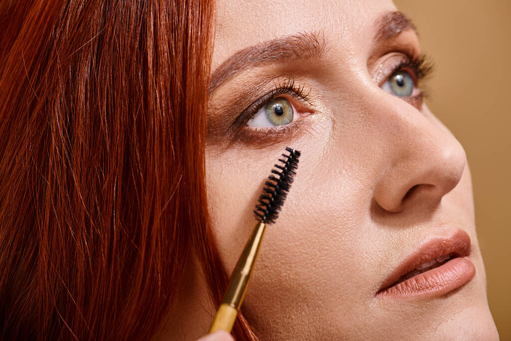 close up vista da mulher ruiva com olhos verdes aplicando rímel no fundo bege, maquiagem - Foto, Imagem