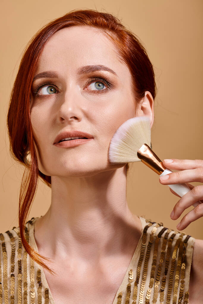 dreamy redhead woman in her 30s applying bronzer with makeup brush on beige background - Photo, Image