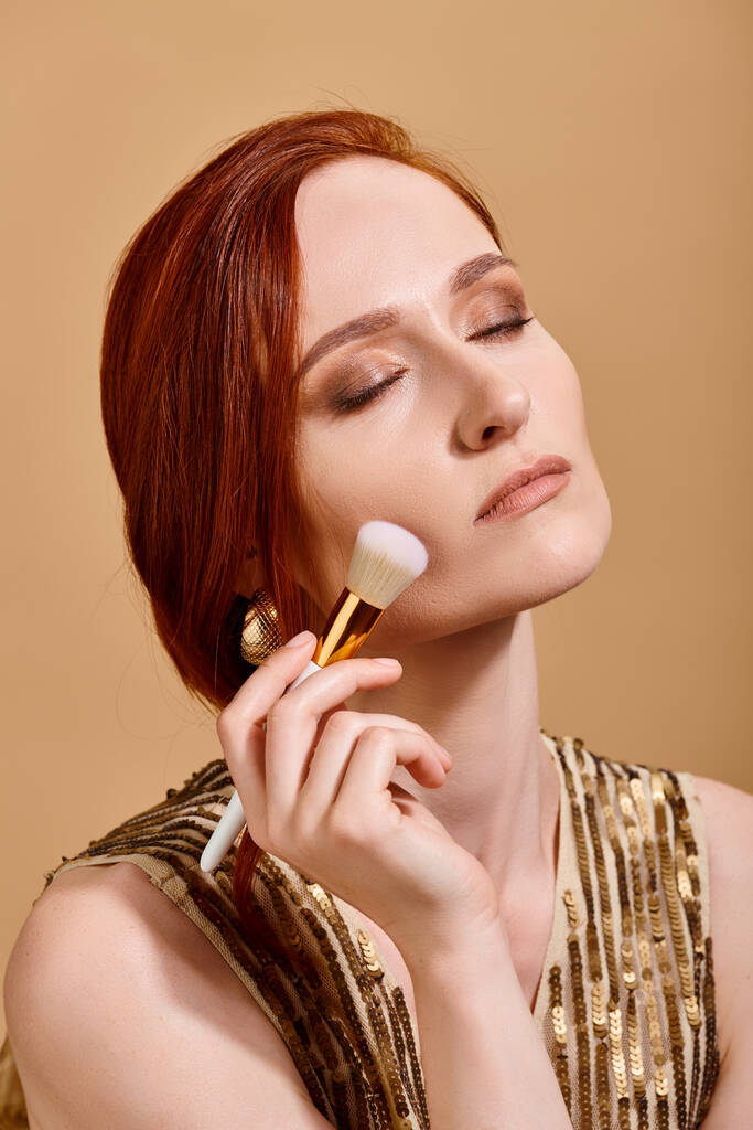 rousse femme en boucle d'oreille en or appliquant fond de teint visage avec pinceau de maquillage sur fond beige - Photo, image