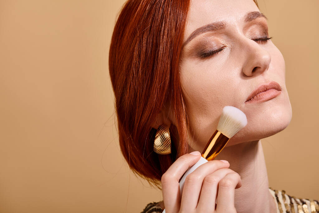 rudowłosa kobieta w złotym kolczyku nakłada podkład pod twarz ze szczotką do makijażu na beżowym tle - Zdjęcie, obraz