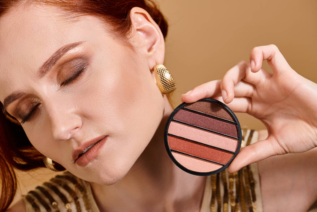 zbliżenie się rudowłosa kobieta trzymając cień do powiek paleta pobliżu twarzy na beżowym tle, makijaż koncepcja - Zdjęcie, obraz