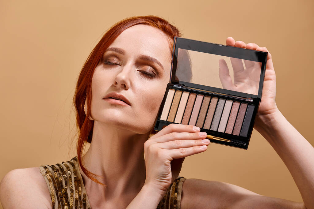 roodharige vrouw met oog schaduw palet in de buurt van gezicht op beige achtergrond, make-up advertentie - Foto, afbeelding