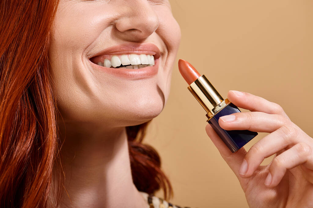 vista recortada de mujer pelirroja sonriendo y aplicando lápiz labial desnudo sobre fondo beige, producto de maquillaje - Foto, imagen