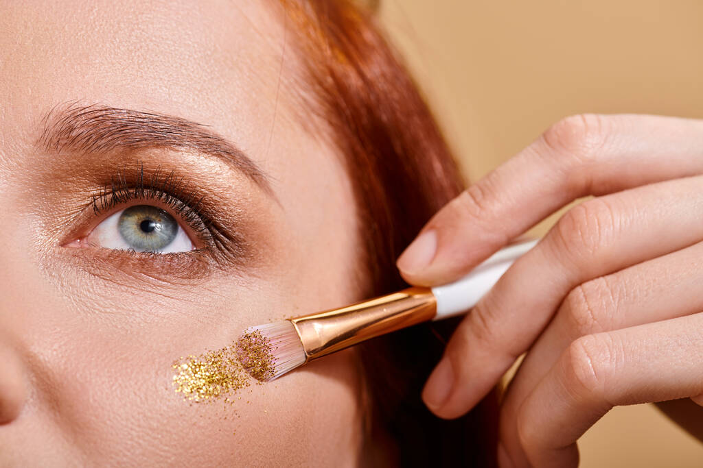 primer plano de mujer pelirroja aplicando brillo brillante en la mejilla con cepillo de maquillaje sobre fondo beige - Foto, imagen