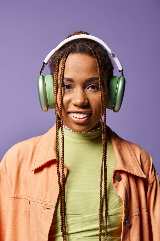 fröhliche afrikanisch-amerikanische Frau in lebendiger Kleidung mit drahtlosen Kopfhörern vor violettem Hintergrund - Foto, Bild