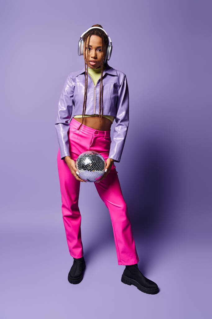 mujer afroamericana joven en elegantes auriculares inalámbricos sosteniendo bola disco sobre fondo púrpura - Foto, imagen