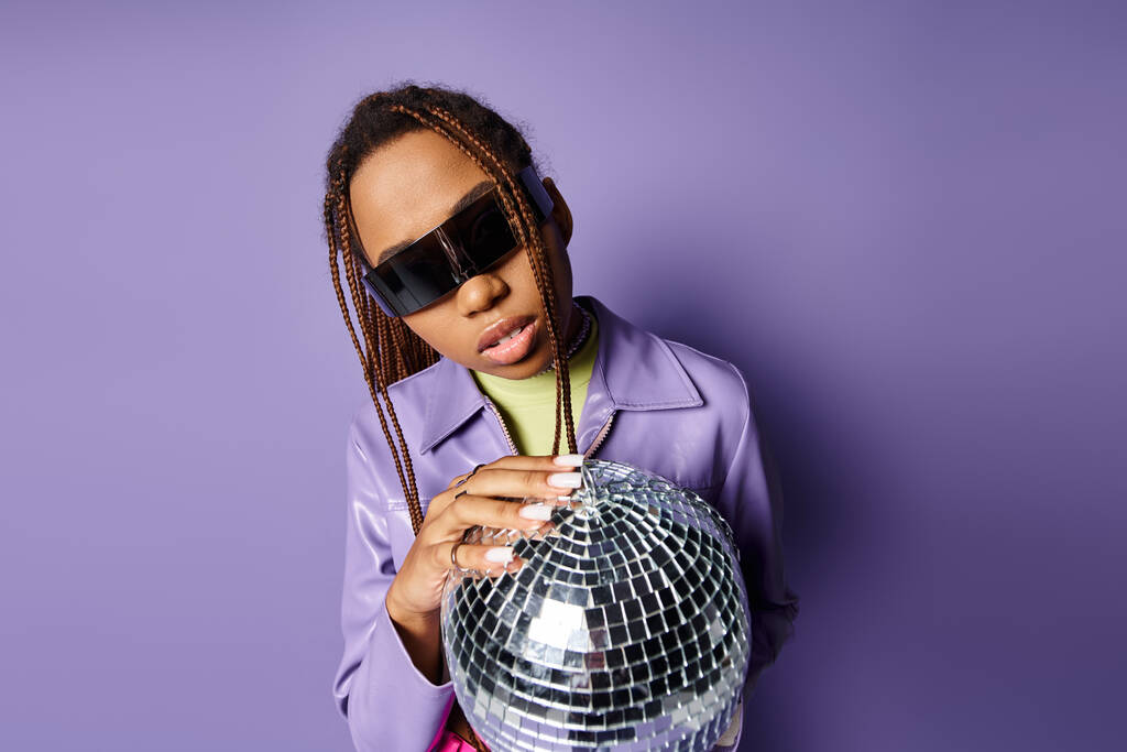 portret van Afrikaanse Amerikaanse vrouw in trendy zonnebril houden disco bal op op paarse achtergrond - Foto, afbeelding