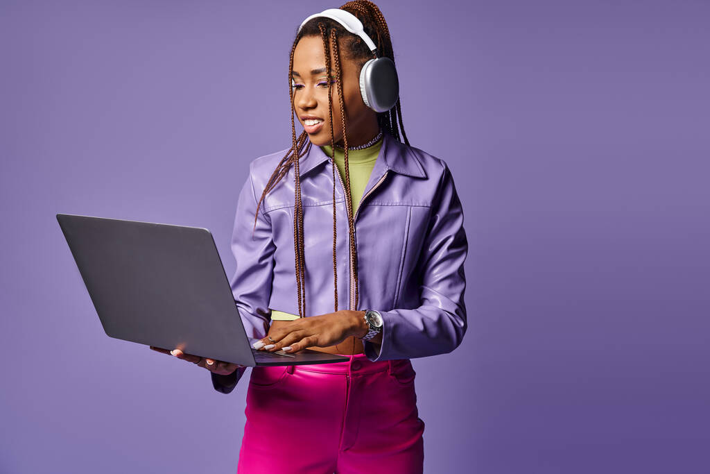 紫色の背景でリモートでラップトップに取り組んでいるヘッドフォンを持つ幸せなアフリカ系アメリカ人女性 - 写真・画像