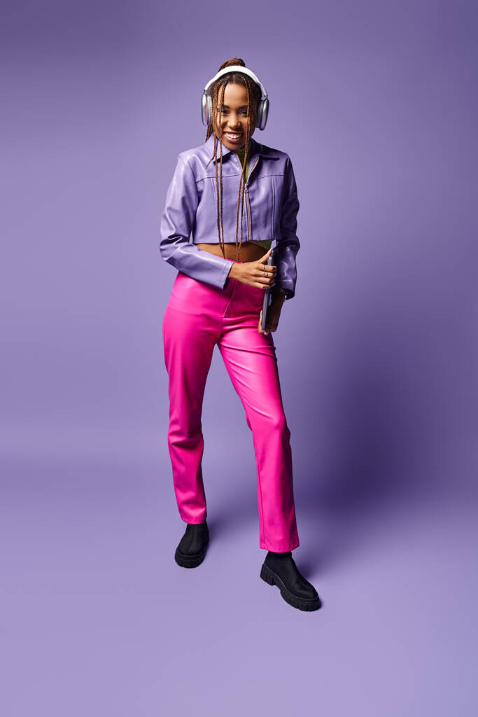 mujer afroamericana feliz con auriculares que sostienen el ordenador portátil y de pie sobre fondo púrpura - Foto, Imagen