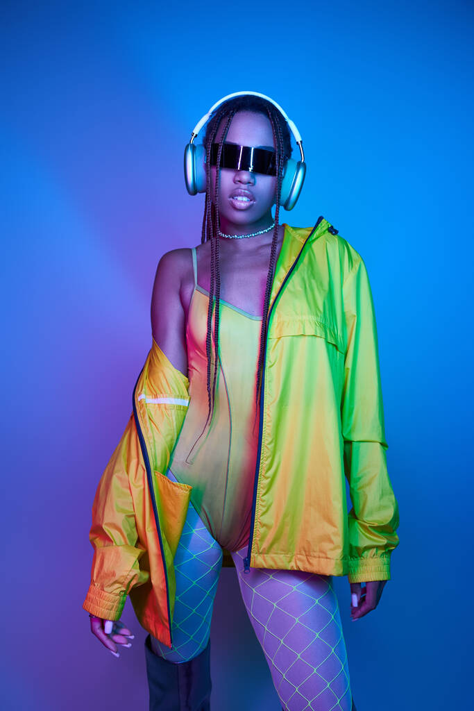 pretty dark-skinned girl in headphones posing in bodysuit and jacket in studio with neon lights - Photo, Image