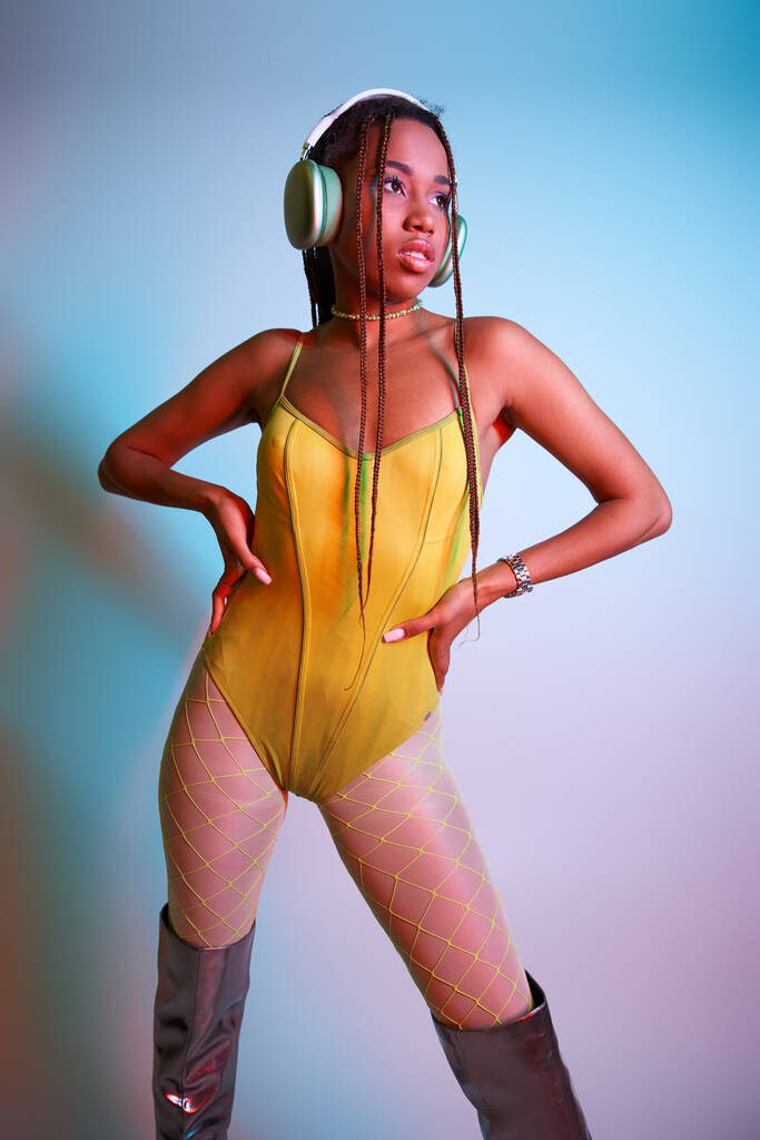 молода афроамериканка в навушниках позує в жовтому костюмі і над коліно чоботи в студії - Фото, зображення