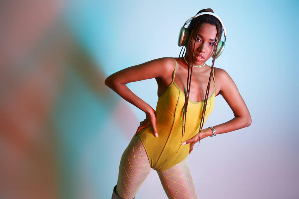 jovem afro-americana em fones de ouvido posando em bodysuit amarelo com as mãos em quadris em estúdio - Foto, Imagem