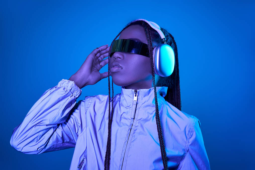 красива темношкіра жінка позує в модних сонцезахисних окулярах з навушниками на синьому з неоновим світлом - Фото, зображення