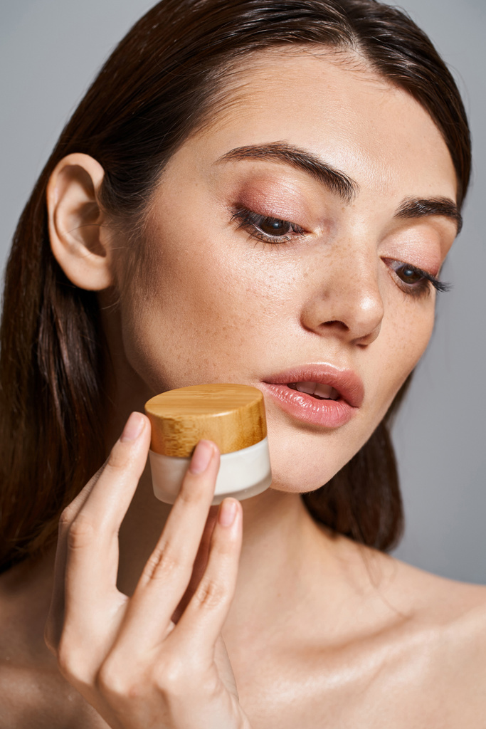 A young Caucasian woman with brunette hair holds a jar of cream to her face, gently applying the luxurious product. - Photo, Image