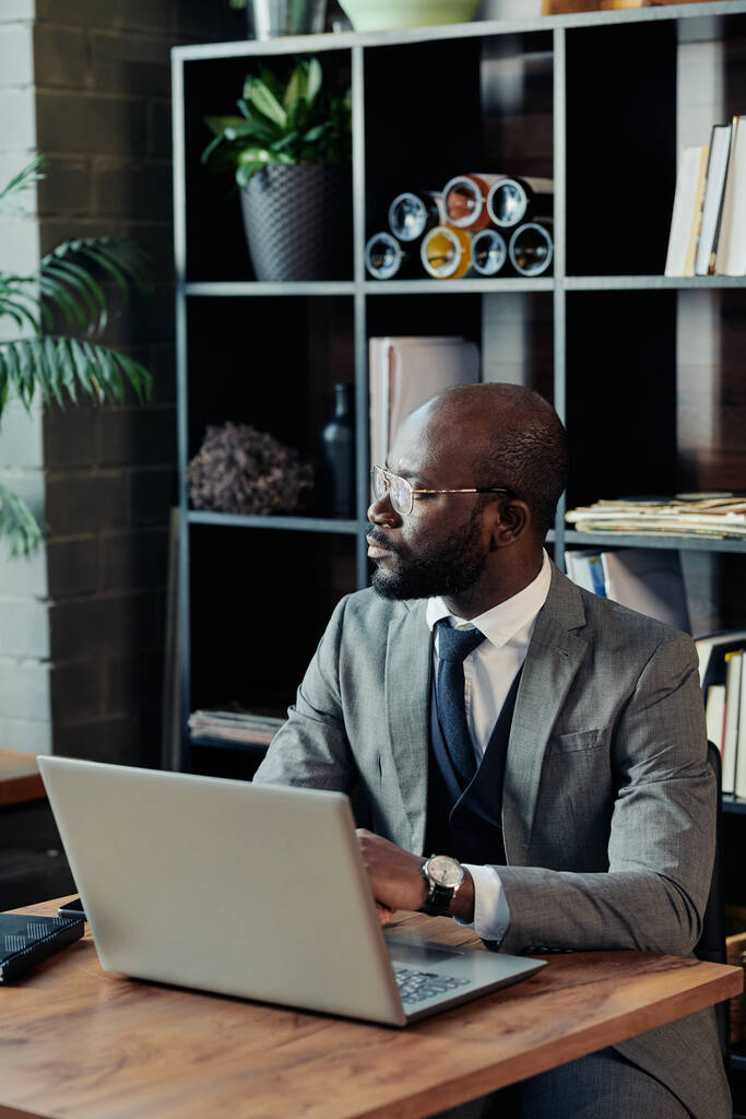 Afrikai férfi öltönyben néz be az ablakon, elmélkedő látvány, miközben dolgozik online laptop kávézóban - Fotó, kép