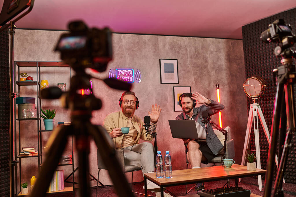 homens barbudos bonitos com fones de ouvido bebendo café e conversando durante podcast, acenando as mãos - Foto, Imagem