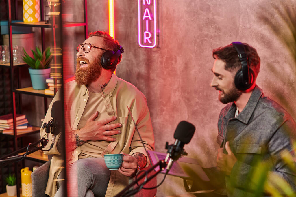 όμορφοι άνδρες σε casual ενδυμασίες ποζάρουν με καφέ και laptop κατά τη διάρκεια της συζήτησης τους στο podcast - Φωτογραφία, εικόνα