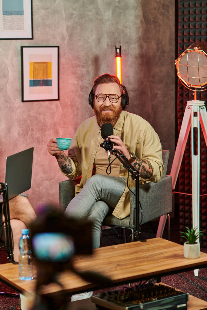 homme gai en tenue décontractée assis avec tasse de café pendant podcast et regardant la caméra - Photo, image