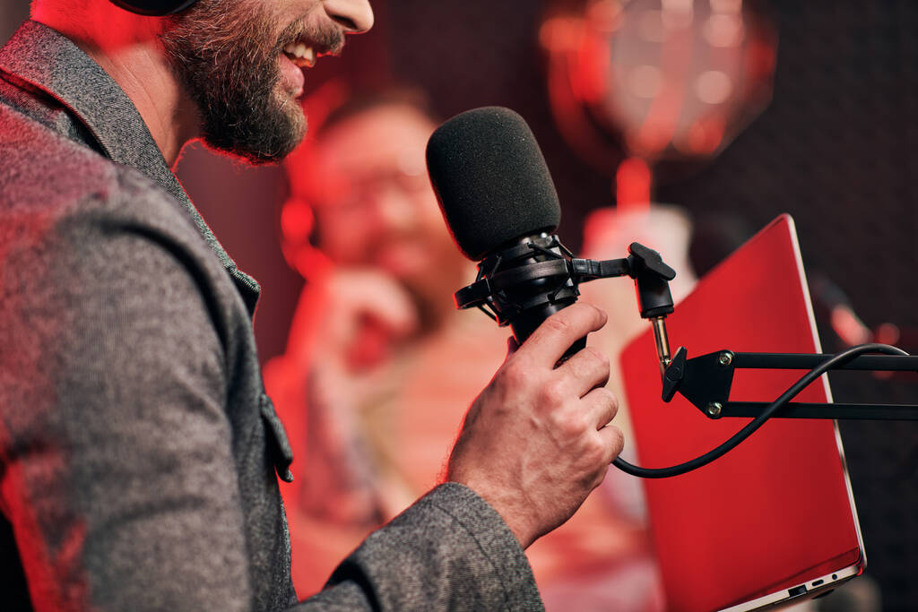 ostřený pohled na vousatého muže s mikrofonem s rozmazaným reportérem na podcastu - Fotografie, Obrázek