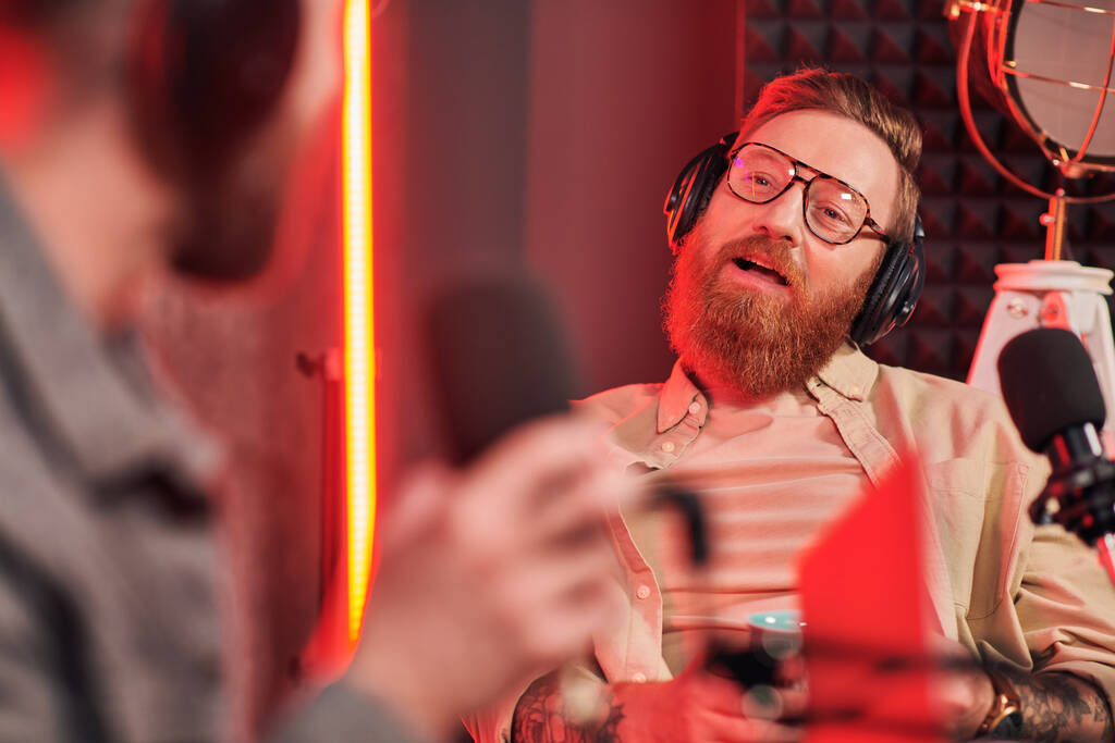 entrevistador barbudo con gafas hablando con su invitado borroso durante su entrevista en podcast - Foto, Imagen