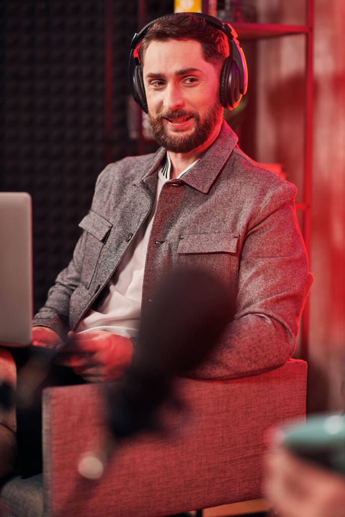 όμορφος γενειοφόρος άνδρας με ακουστικά σε casual outfit κάθεται κατά τη διάρκεια podcast ενώ στο στούντιο - Φωτογραφία, εικόνα