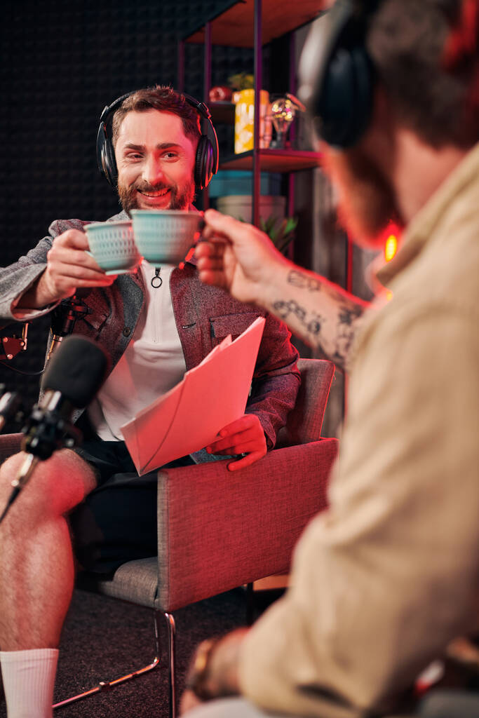 vrolijk bebaarde man met koffiekop in zijn hand op zoek naar papierwerk naast zijn podcaster - Foto, afbeelding