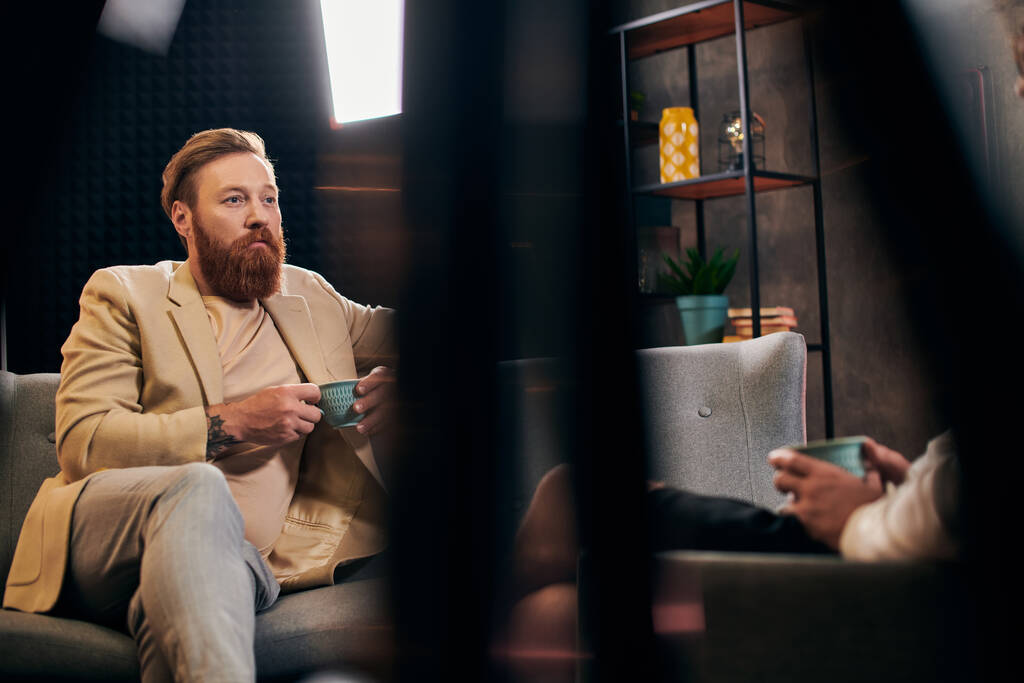 hombre barbudo guapo con pelo rojo en ropa elegante sentado al lado de su entrevistador en el estudio - Foto, imagen
