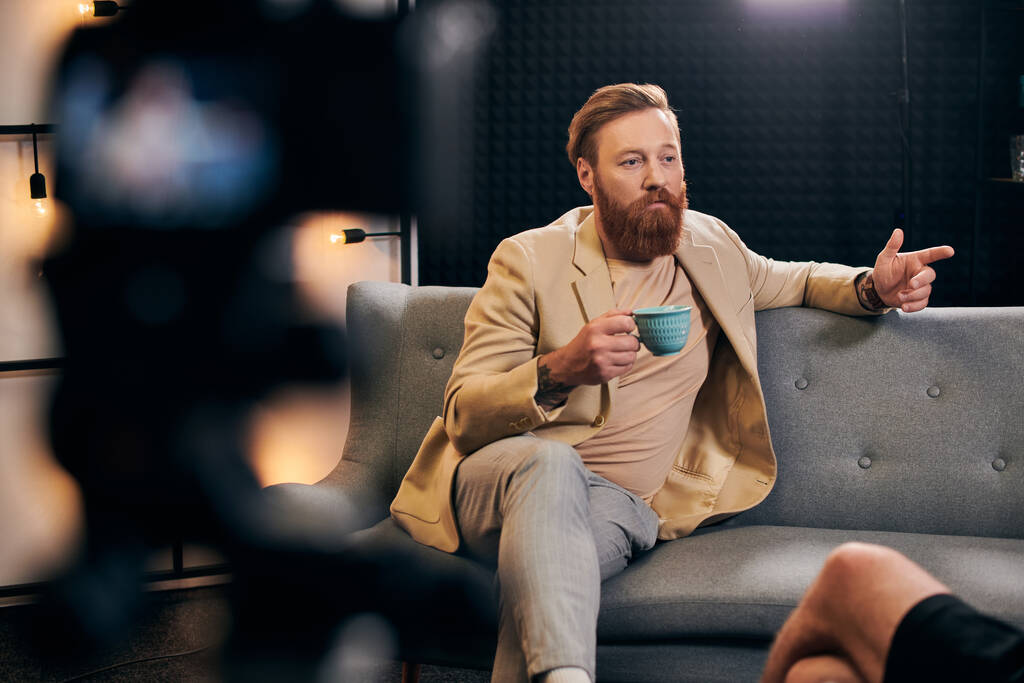 jóképű szakállas férfi, vörös hajjal, elegáns ruhában, aki a stúdióban ül az interjúalany mellett. - Fotó, kép