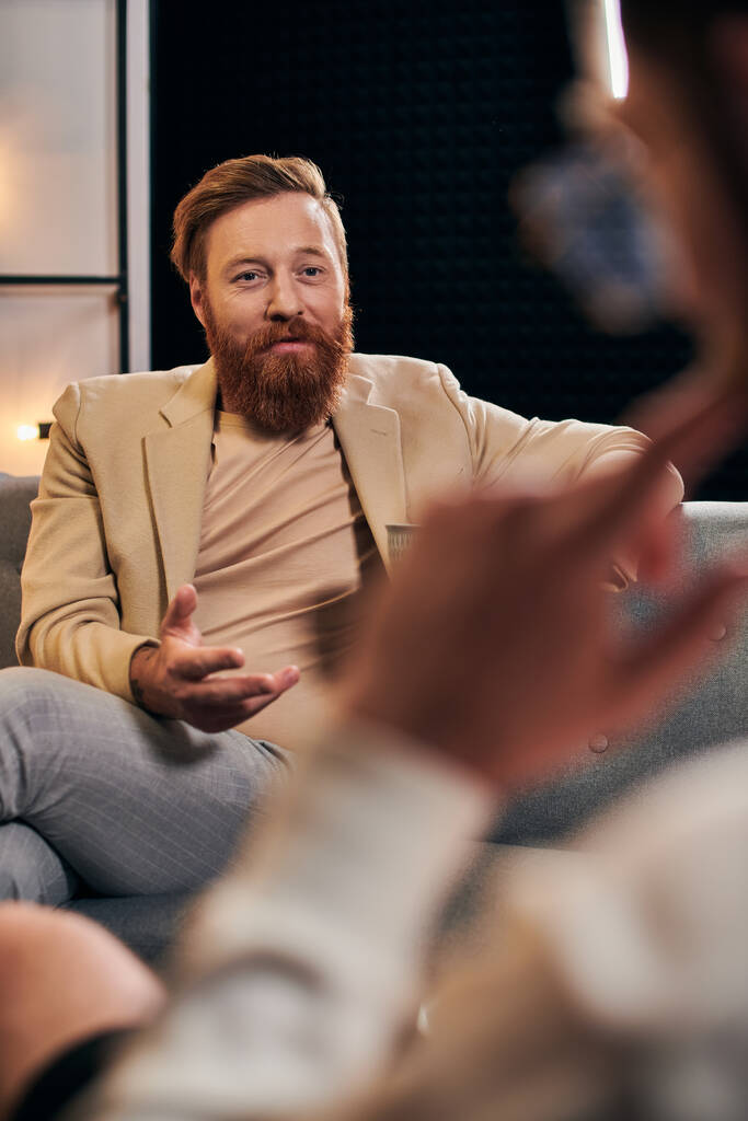 jóképű szakállas férfi, vörös hajjal, elegáns ruhákban, a riporter mellett ül a stúdióban. - Fotó, kép
