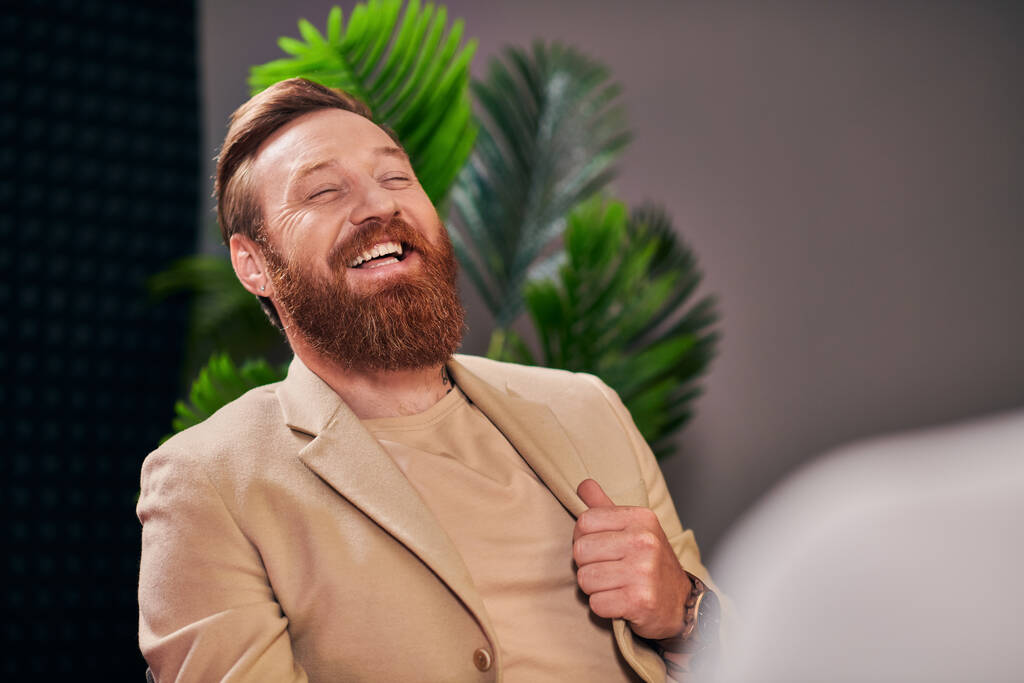 guapo hombre alegre con barba en elegante atuendo riendo durante la entrevista mientras está en el estudio - Foto, imagen