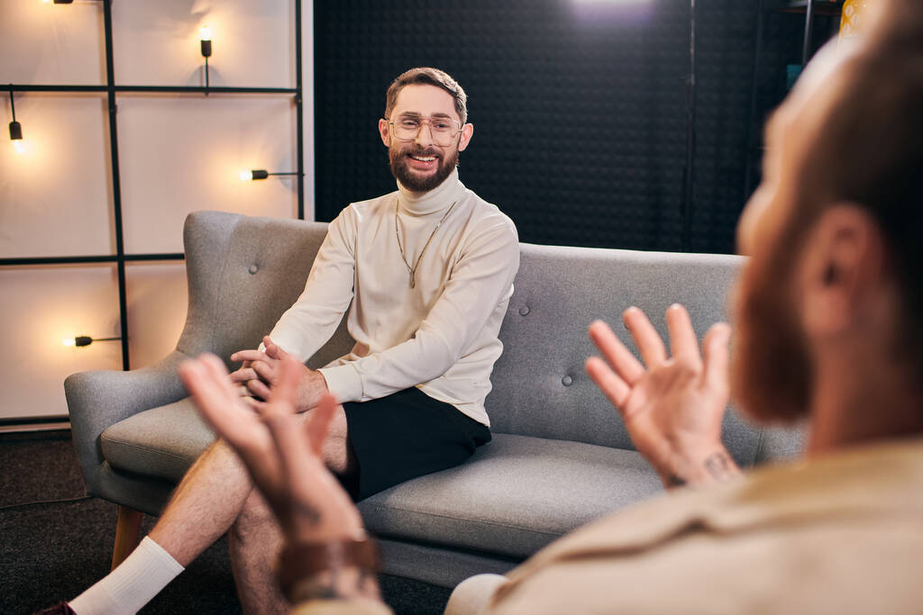 vrolijk baard mannen in casual outfit glimlachen en kijken naar zijn interviewer terwijl zitten in de studio - Foto, afbeelding